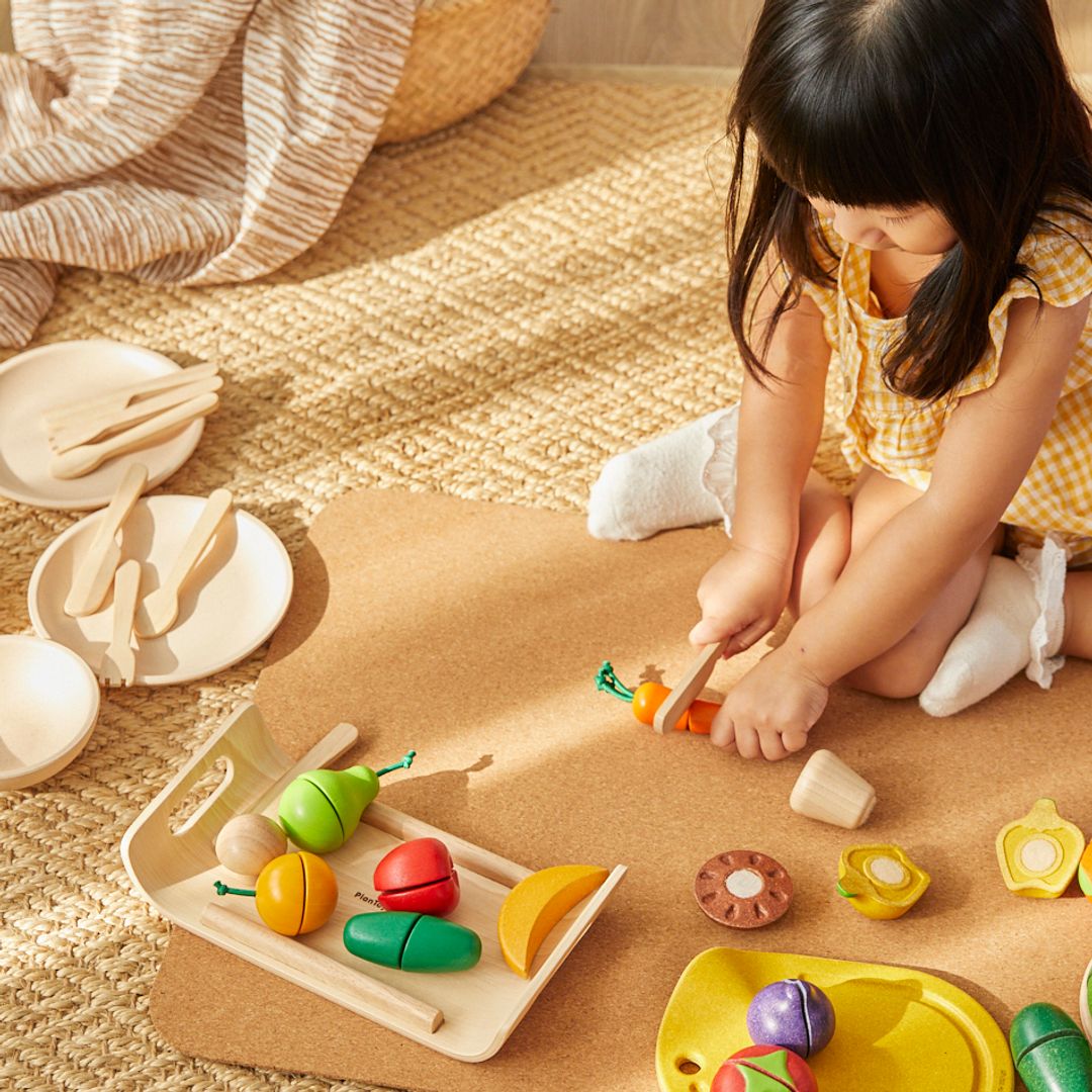 Overhead view of a little girl practicing fine motor skills by slicing the wooden carrot with the PlanToys play knife.