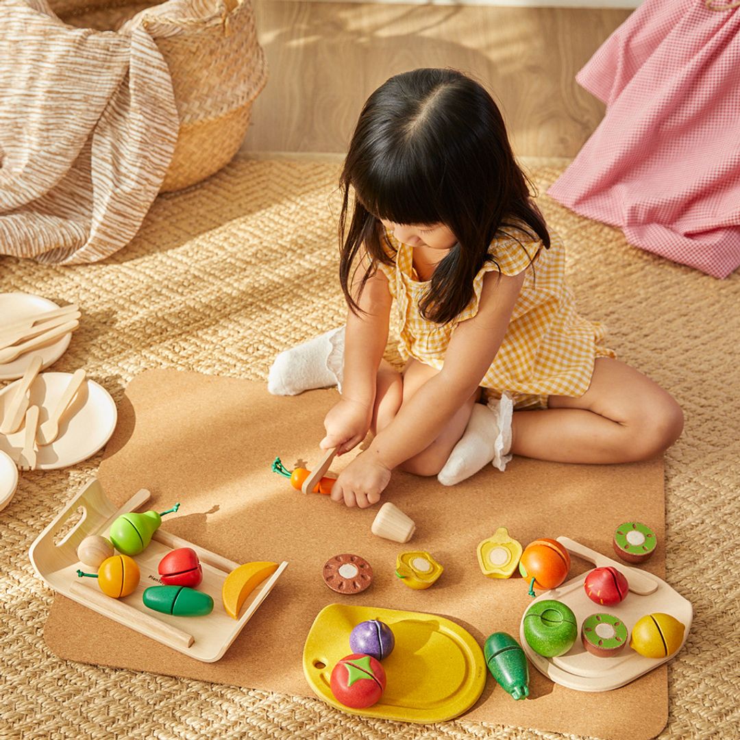 A toddler girl enjoying imaginative play with multiple PlanToys wooden food sets on a play mat.