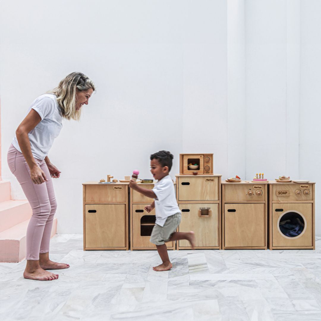 A mother and son laughing and playing together near the durable, earth-friendly PlanToys wooden play kitchen collection.