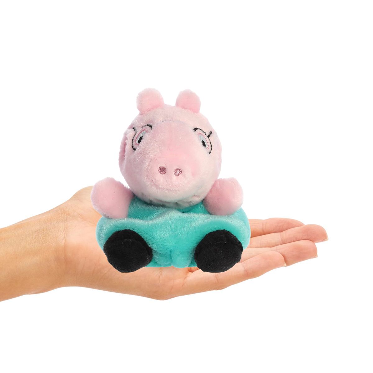 Small plush toy of a pink pig in a green shirt held in a hand on a white background