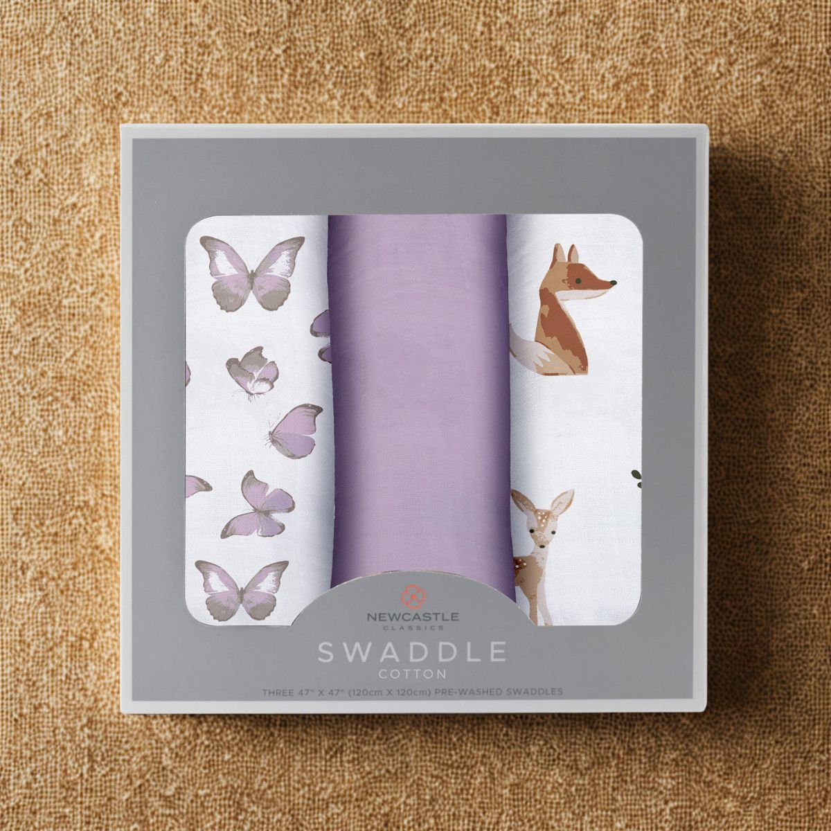 Straight top-down view of the packaged Mountain Meadow baby swaddle blankets inside a grey Newcastle Classics 3-pack box resting on a textured cork surface.