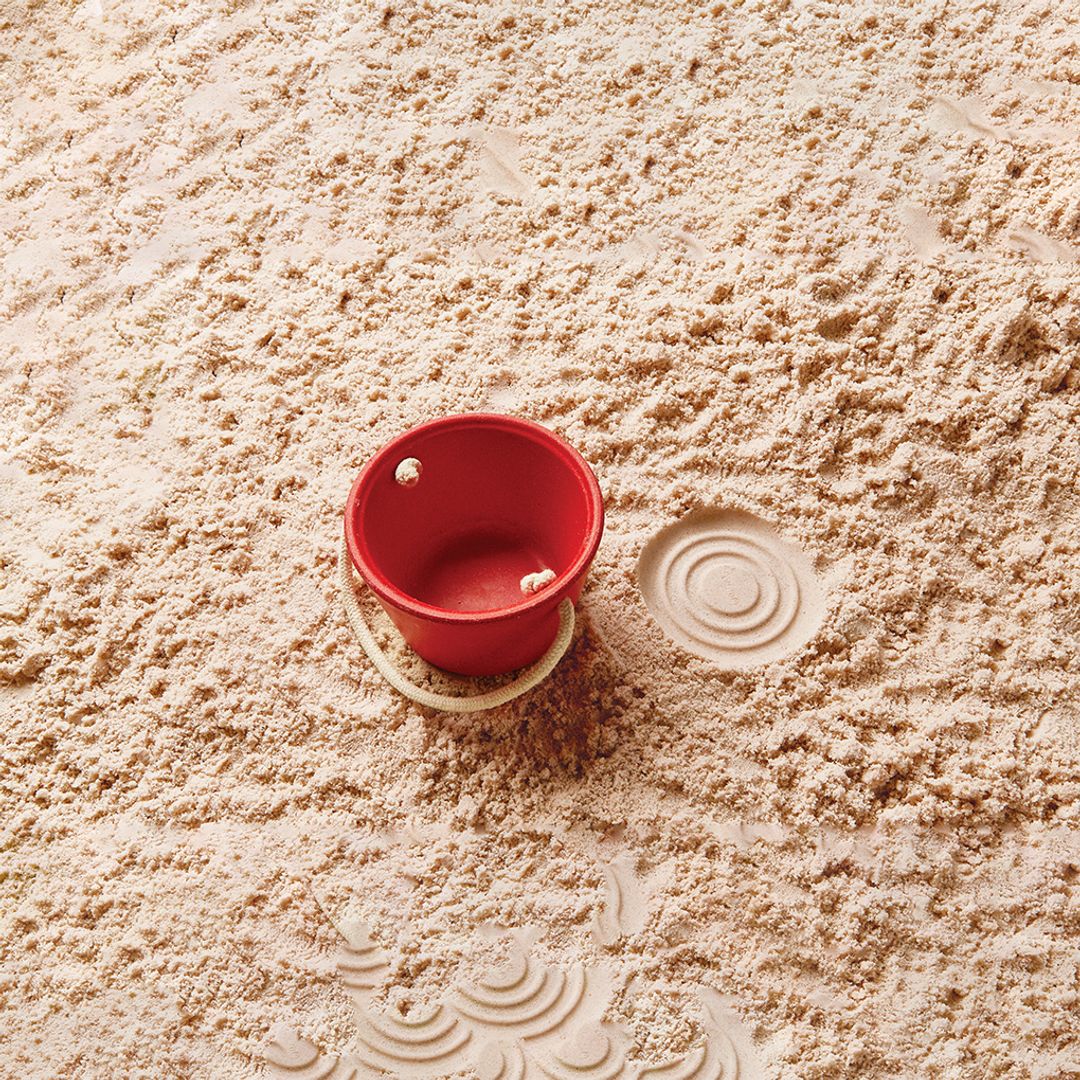 Red PlanToys wooden sand bucket resting on sand next to a circular bucket imprint.