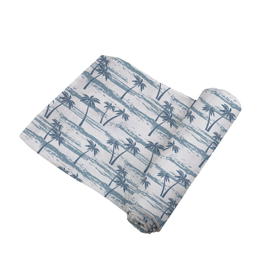 Angled view of the white swaddle blanket with blue palm trees and wave lines partially unrolled to show its soft texture.
