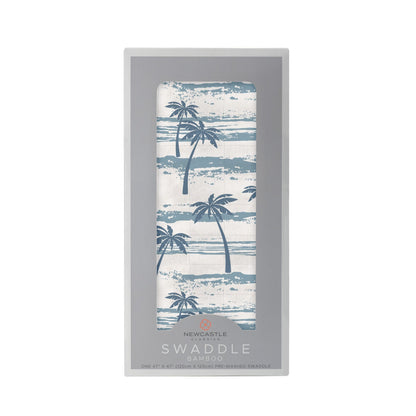 Straight front view of the packaged Ocean Palm Trees baby swaddle blanket inside a grey Newcastle Classics display box.