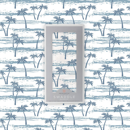 Flat lay of the Newcastle Classics Ocean Palm Trees bamboo muslin swaddle blanket shown with its branded grey packaging box in the center against a matching palm tree pattern background.