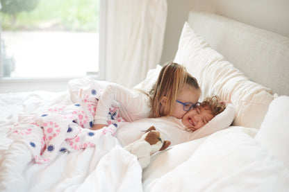 Lifestyle shot of an older sister cuddling a baby wrapped in the pink and indigo floral swaddle blanket on a white bed.