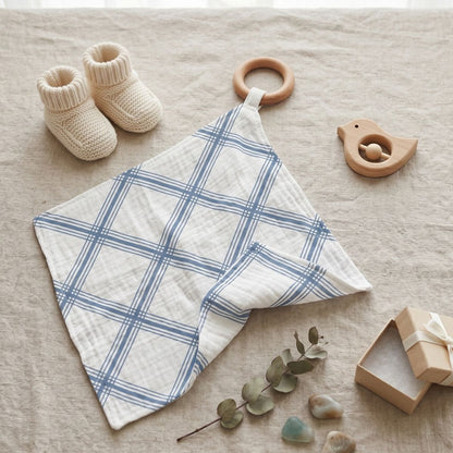 The blue plaid muslin blankie teether displayed flat on a beige fabric surface next to knit baby booties and a wooden bird toy.