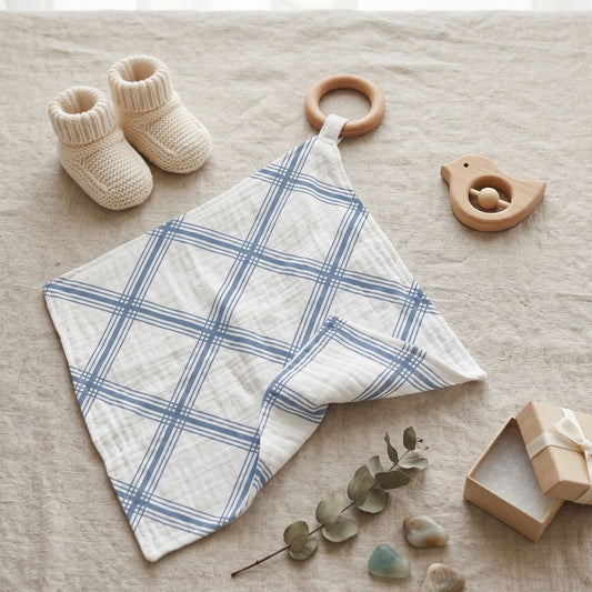 The blue plaid muslin blankie teether displayed flat on a beige fabric surface next to knit baby booties and a wooden bird toy.