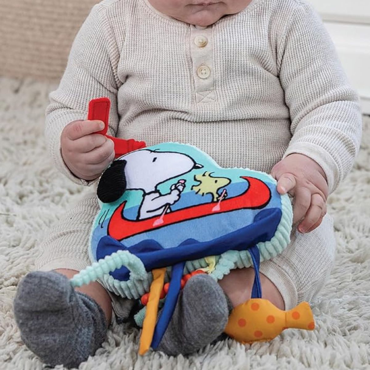 A baby sitting down on a soft blanket while happily holding and playing with the colorful Snoopy plush sensory toy.
