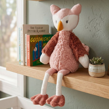 Happy Horse Owl Olivia plush toy sitting on a floating wooden nursery shelf with her legs dangling next to classic children's books.
