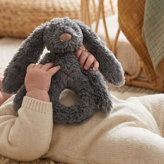 A baby resting and holding onto the Happy Horse Rabbit Richie deep grey plush rattle with both hands.