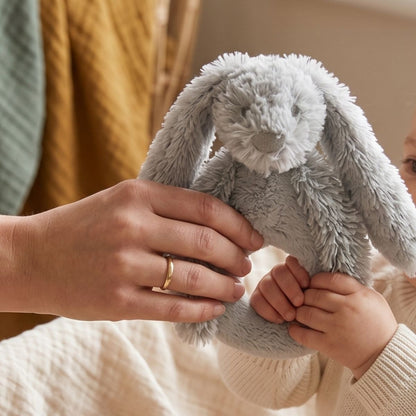 An adult's hands helping a baby's tiny hands hold the Happy Horse Rabbit Richie classic grey plush rattle.