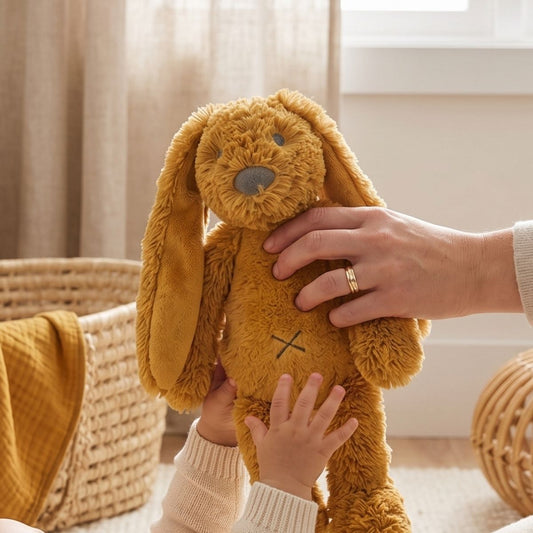 An adult's hands supporting the Happy Horse Rabbit Richie ochre plush toy as a baby interacts with it in a cozy nursery setting.