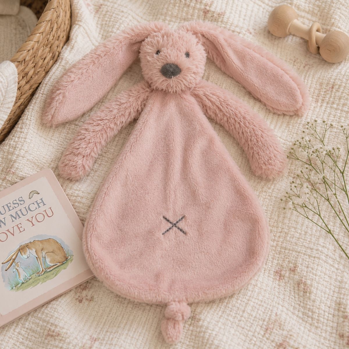 Happy Horse Rabbit Richie old pink plush lovey resting flat on a cozy knit blanket inside a nursery setting next to a baby book.