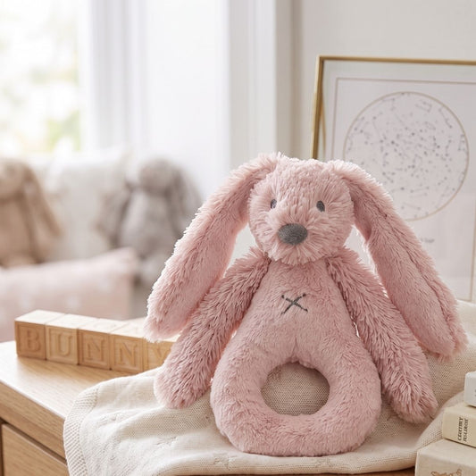 Happy Horse Rabbit Richie old pink plush rattle sitting upright on a soft blanket in a cozy, bright nursery setting.