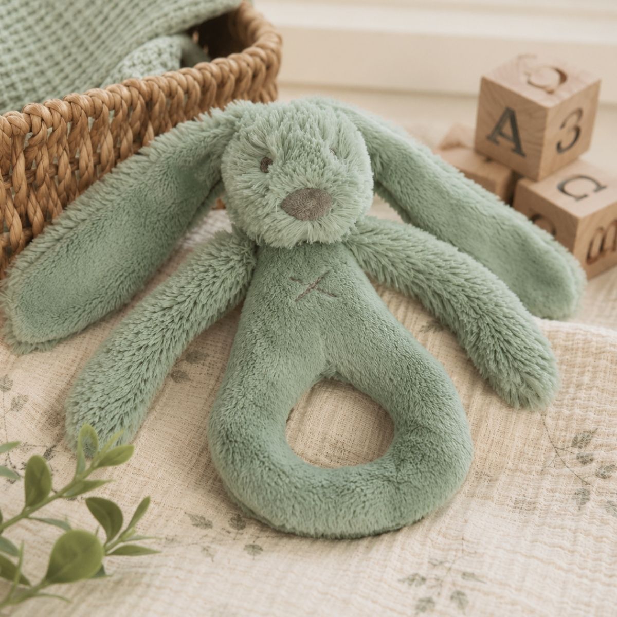 Happy Horse Rabbit Richie green plush rattle resting on a light blanket next to wooden alphabet blocks and a storage basket.