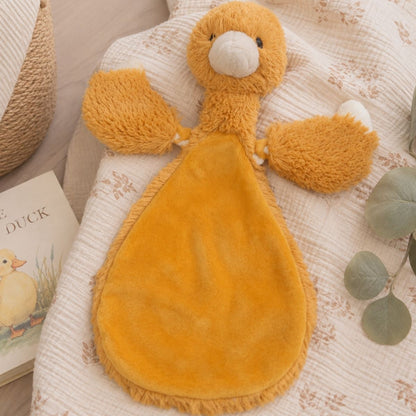 Happy Horse Duck Twine ochre plush lovey resting flat on a cozy blanket inside a nursery setting next to a decorative eucalyptus branch.