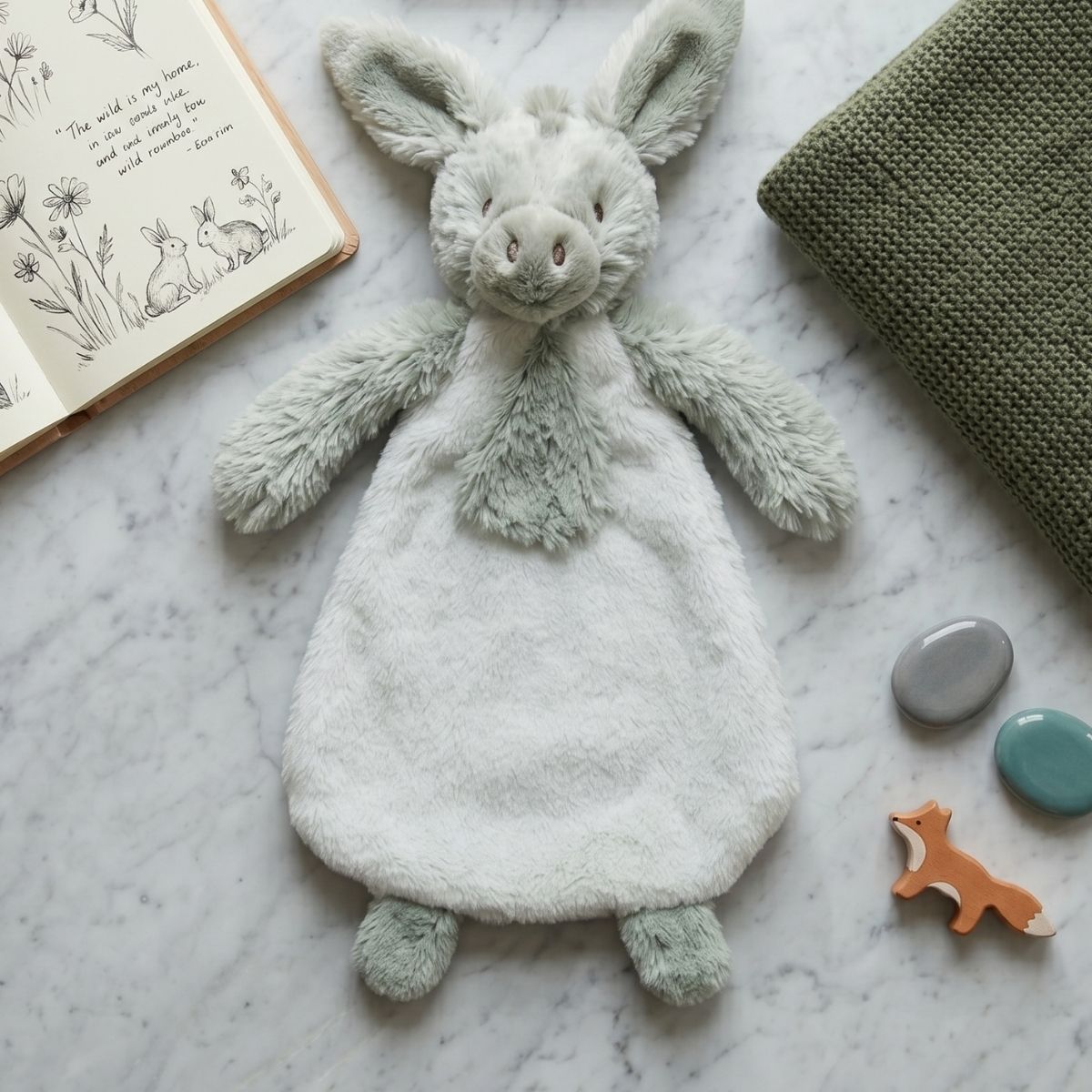 Happy Horse Donkey Diego plush lovey laid flat on a gray surface next to an open baby book and a green blanket.