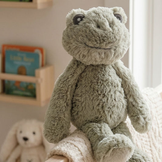Happy Horse Frog Flex plush toy sitting on a cozy blanket on a nursery chair next to a floating wooden bookshelf.