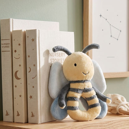 Happy Horse Bee Benja plush rattle resting against a stack of white nursery books on a wooden shelf.