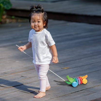 A smiling toddler girl walking on a wooden deck while pulling her vibrant PlanToys rainbow alligator.