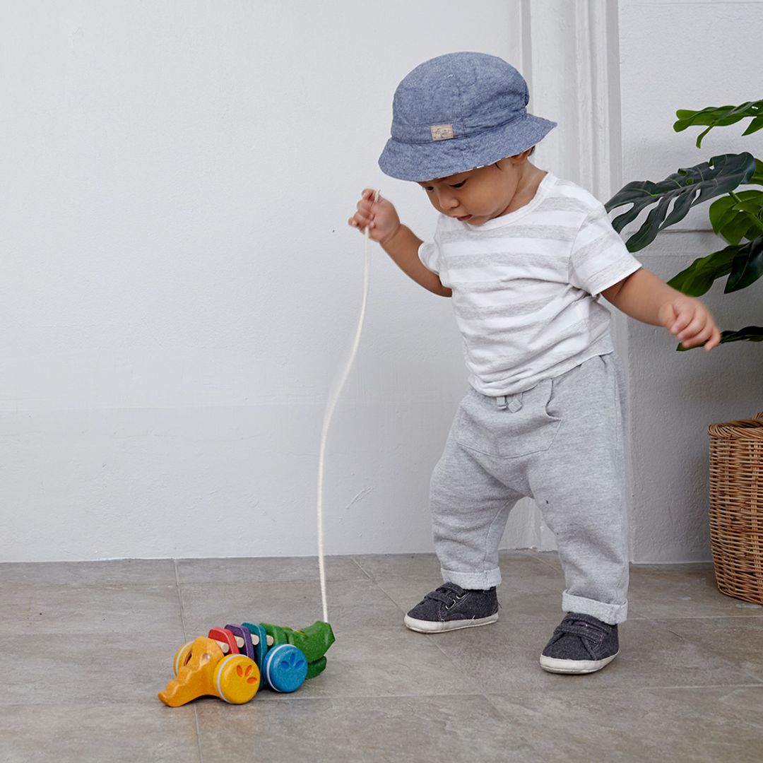 A young boy in a hat practicing his walking skills indoors by towing the earth-friendly PlanToys alligator toy.