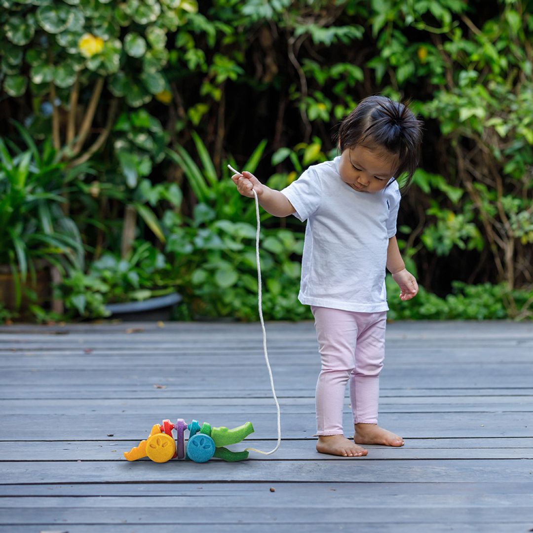 A toddler girl turning around to look at her colorful PlanToys wooden pull-along alligator during outdoor play.