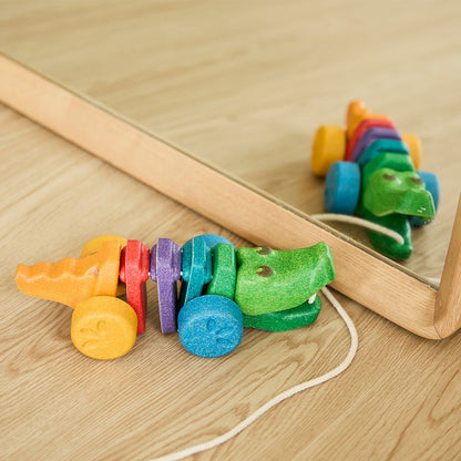 The chemical-free PlanToys rainbow alligator toy resting on a wooden floor next to a floor mirror.