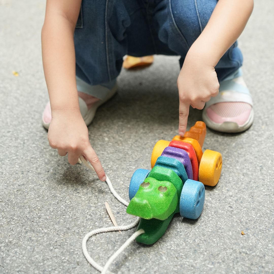 Close-up of a child's hands pointing at the head of the eco-friendly PlanToys rainbow alligator pull toy.