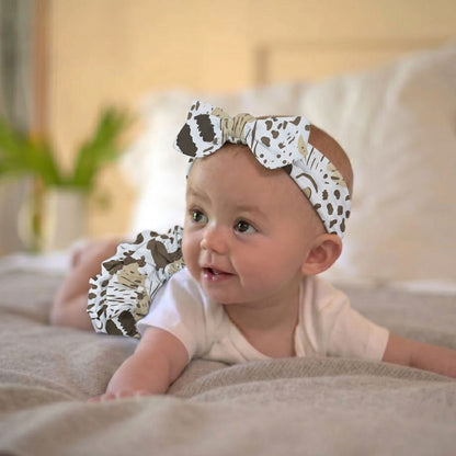 Baby girl wearing the Newcastle Classics On The Savannah headband and matching ruffled bloomers smiling while laying on a bed.