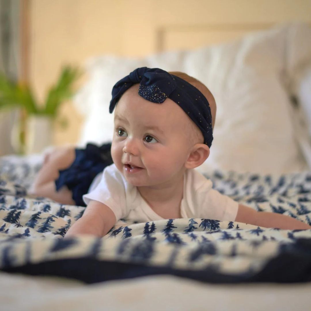 Baby girl wearing a dark blue Newcastle Classics headband while playing on a patterned blanket.