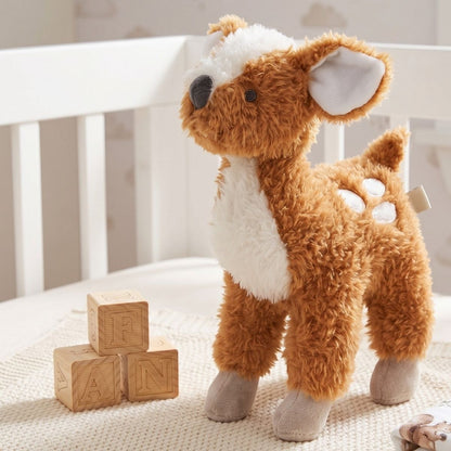 Happy Horse Deer Doe plush fawn standing on a textured blanket next to wooden playing blocks near a white nursery crib.