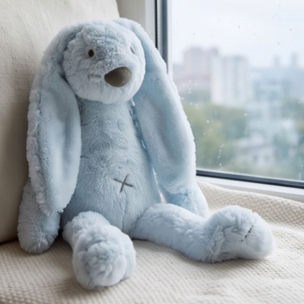 Happy Horse Rabbit Richie blue plush toy sitting upright on a cozy indoor chair next to a window overlooking a cityscape.
