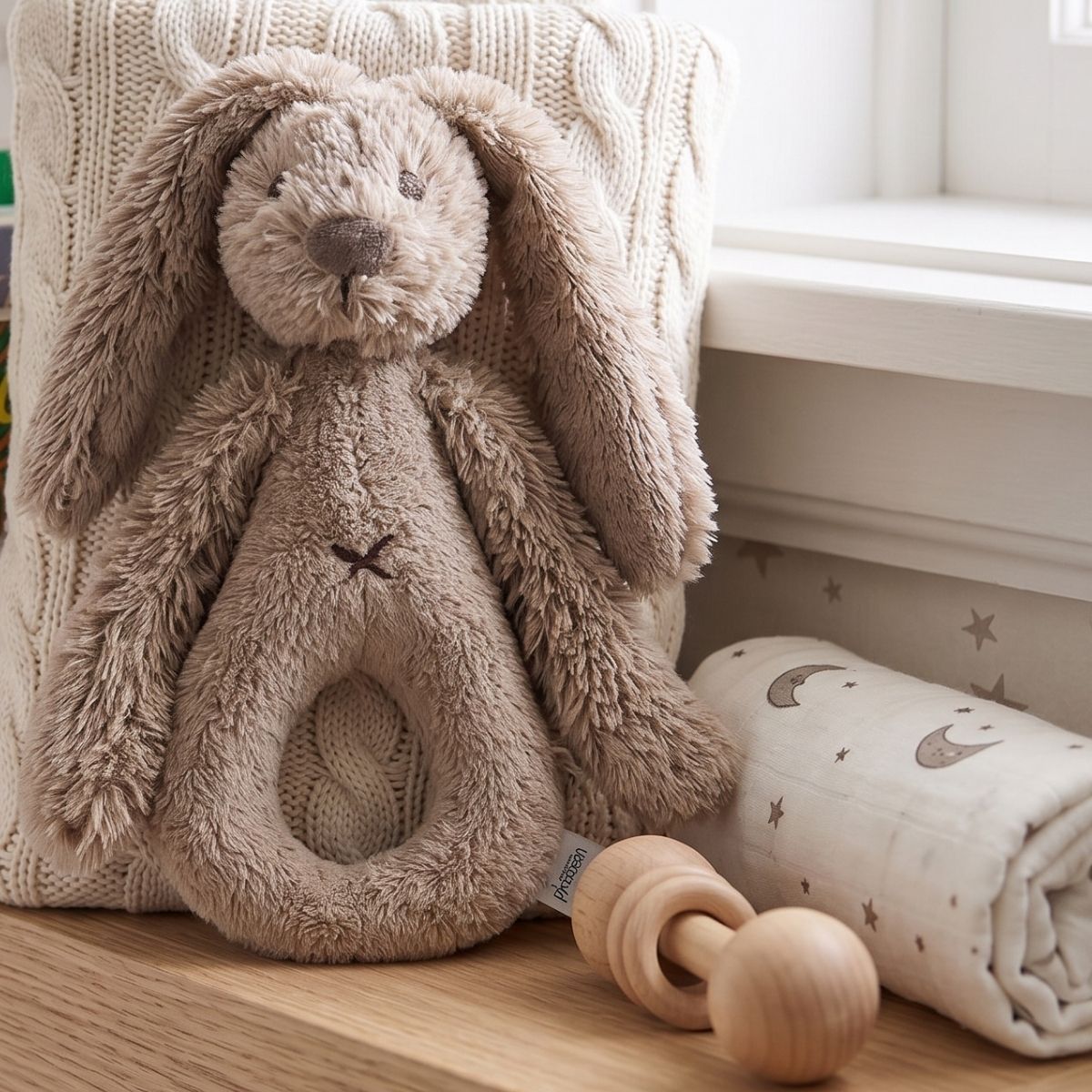 Happy Horse Rabbit Richie clay plush rattle sitting upright on a wooden shelf next to a rolled baby blanket and wooden teething toy.