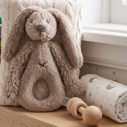 Happy Horse Rabbit Richie clay plush rattle sitting upright on a wooden shelf next to a rolled baby blanket and wooden teething toy.