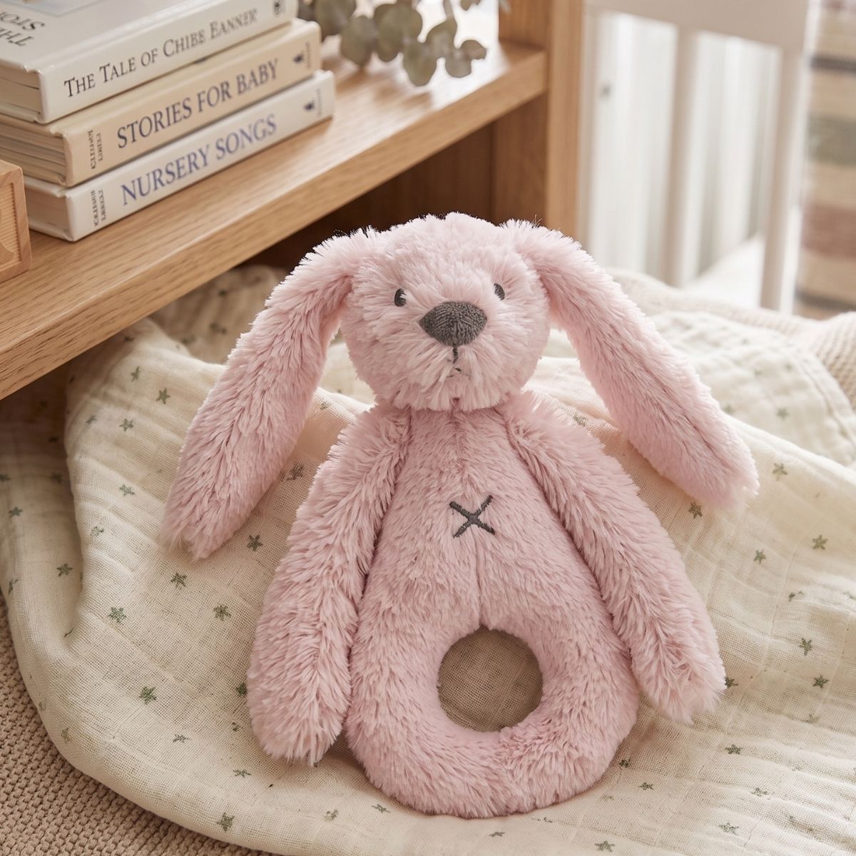 Happy Horse Rabbit Richie pink plush rattle lying on a soft baby blanket next to a wooden shelf with nursery books.