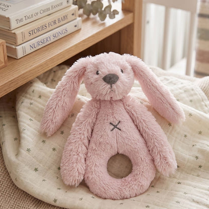 Happy Horse Rabbit Richie pink plush rattle lying on a soft baby blanket next to a wooden shelf with nursery books.