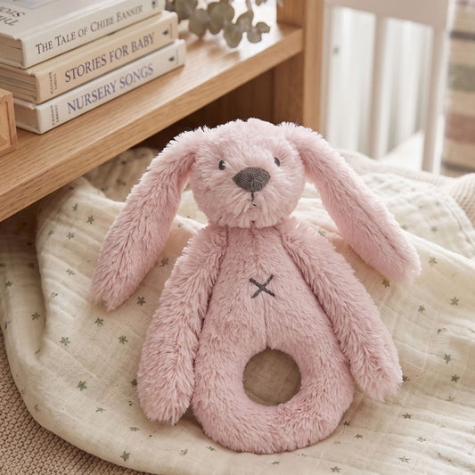 Happy Horse Rabbit Richie pink plush rattle lying on a soft baby blanket next to a wooden shelf with nursery books.