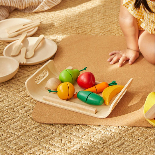 A toddler sitting next to the PlanToys wooden serving tray filled with colorful, sliceable play food.