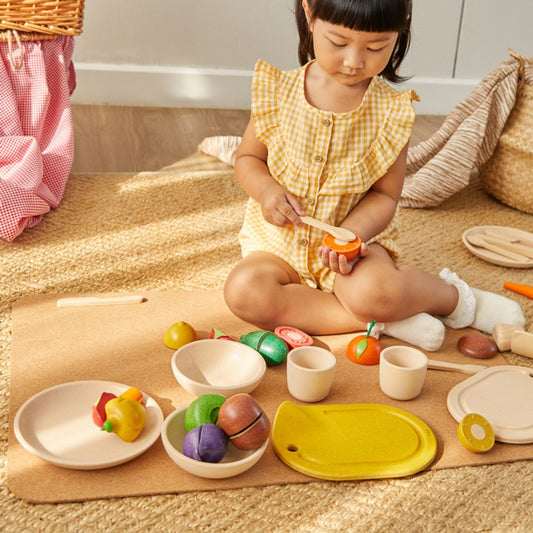 A toddler girl enjoying imaginative play with her PlanToys wooden dishes and play food on a woven floor mat.