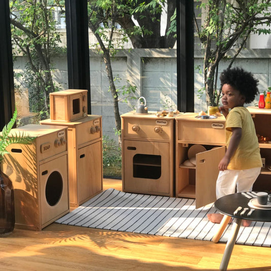 A toddler boy enjoying imaginative pretend play in a full PlanToys sustainable wooden kitchen setup.