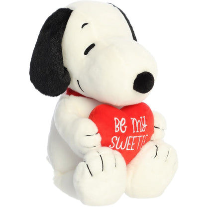 Aurora 11-inch Peanuts Snoopy Be My Sweetie plush toy sitting at an angle holding a vibrant red plush heart that reads "Be My Sweetie" in white cursive script.