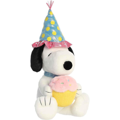 Angled front view of the 12-inch Aurora Peanuts Birthday Snoopy stuffed animal sitting down wearing a party hat and holding a cupcake.