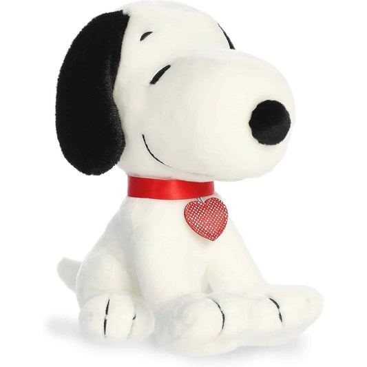 Aurora 9-inch Peanuts Seated Snoopy plush toy sitting at an angle showing its white fur, closed happy eyes, red collar with a sparkling red heart tag, and black floppy ears.