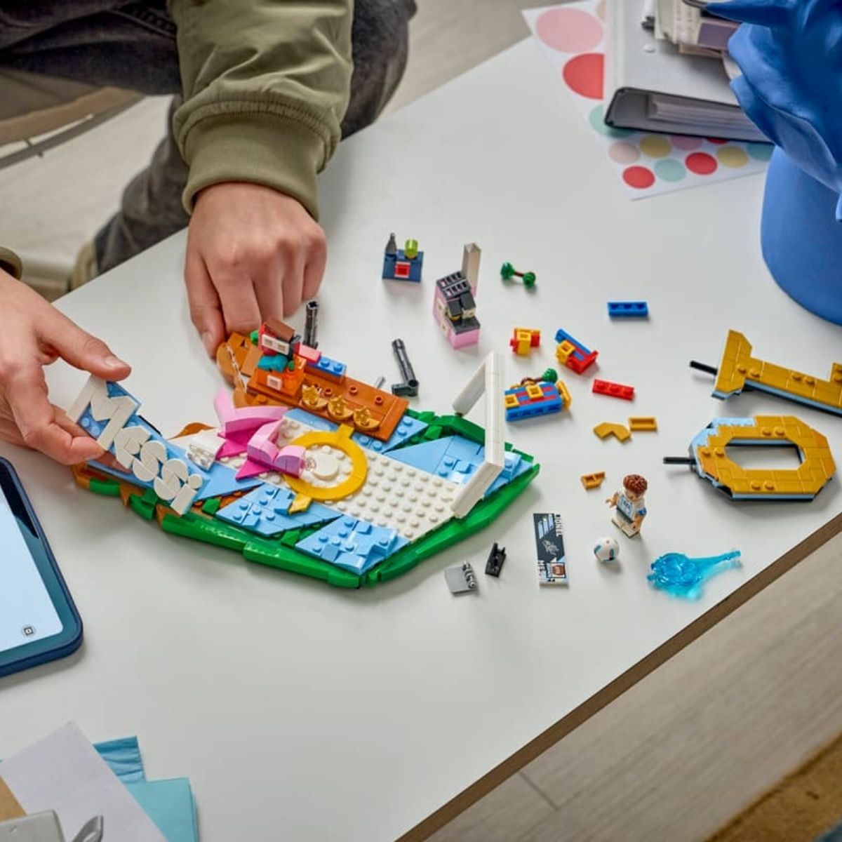 A pair of hands building the 500-piece LEGO® Lionel Messi soccer highlights set with scattered pieces on a table.