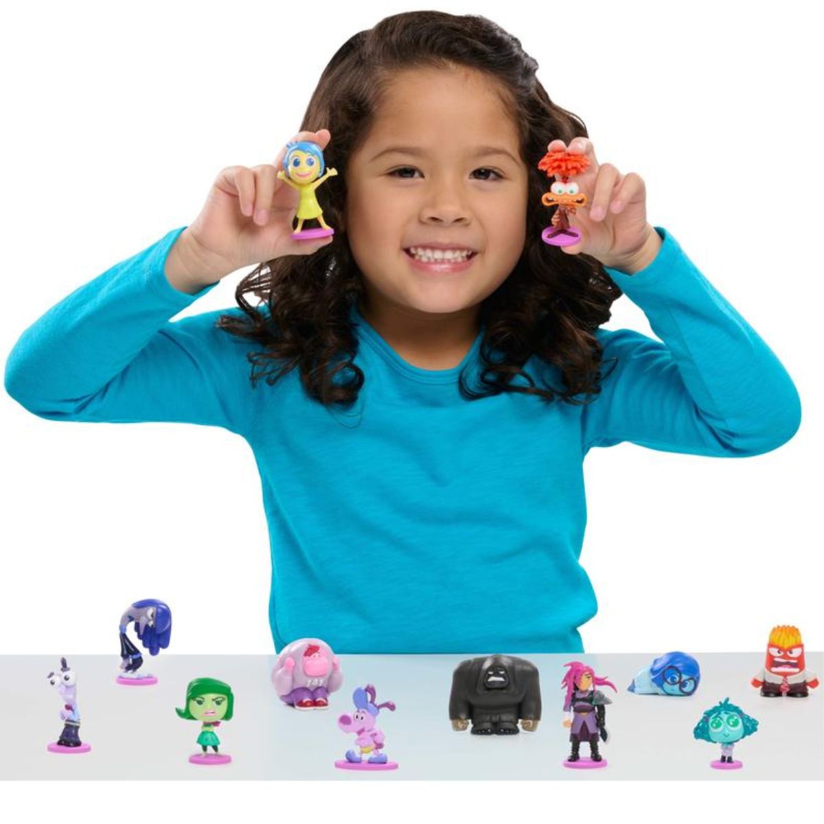 A young girl smiling while showing off two colorful Inside Out 2 mini emotion figures held up next to her ears.