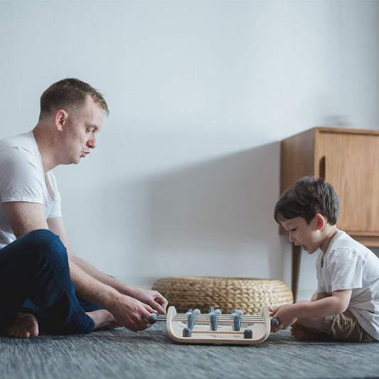 A father and son sitting on the floor setting up the sustainable PlanToys wooden foosball game together.