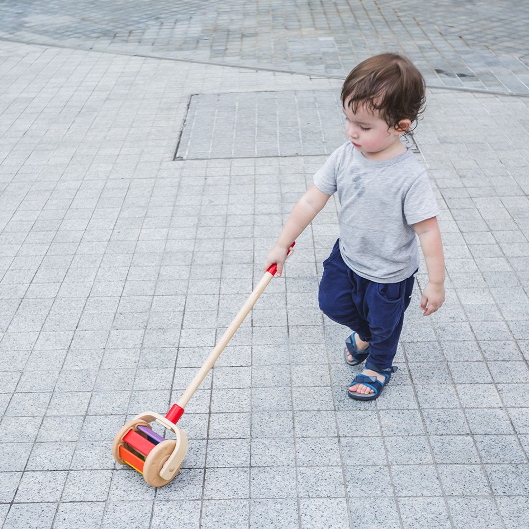 A toddler boy developing gross motor skills by pushing the colorful PlanToys wooden walking toy outside on a paved path.