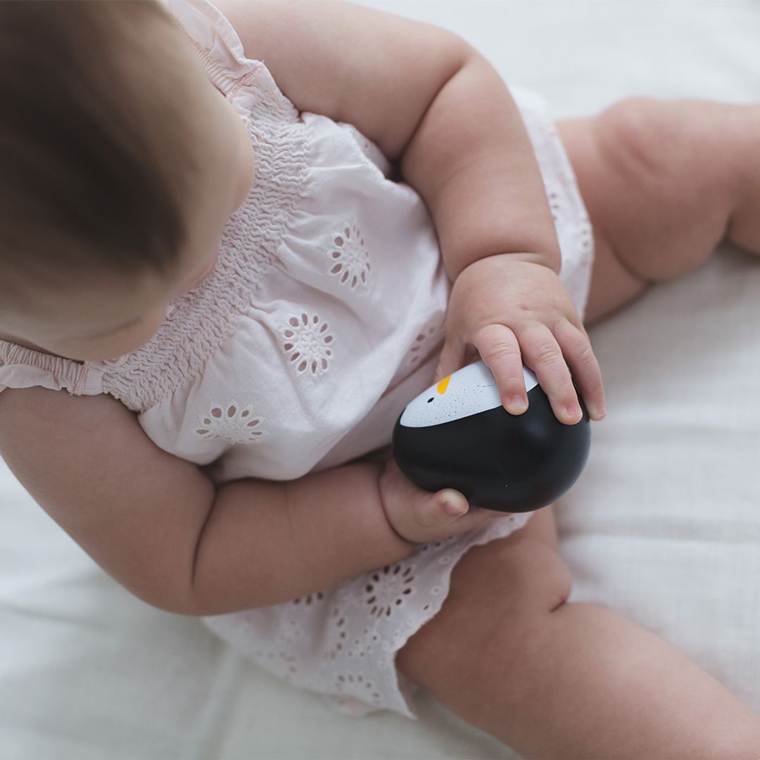 A baby sitting in a white onesie holding the smooth, rounded PlanToys wooden penguin wobbler.