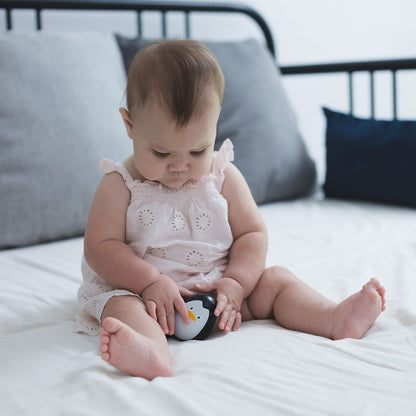 A baby sitting on a white bed with grey pillows looking down at the earth-friendly PlanToys penguin toy.